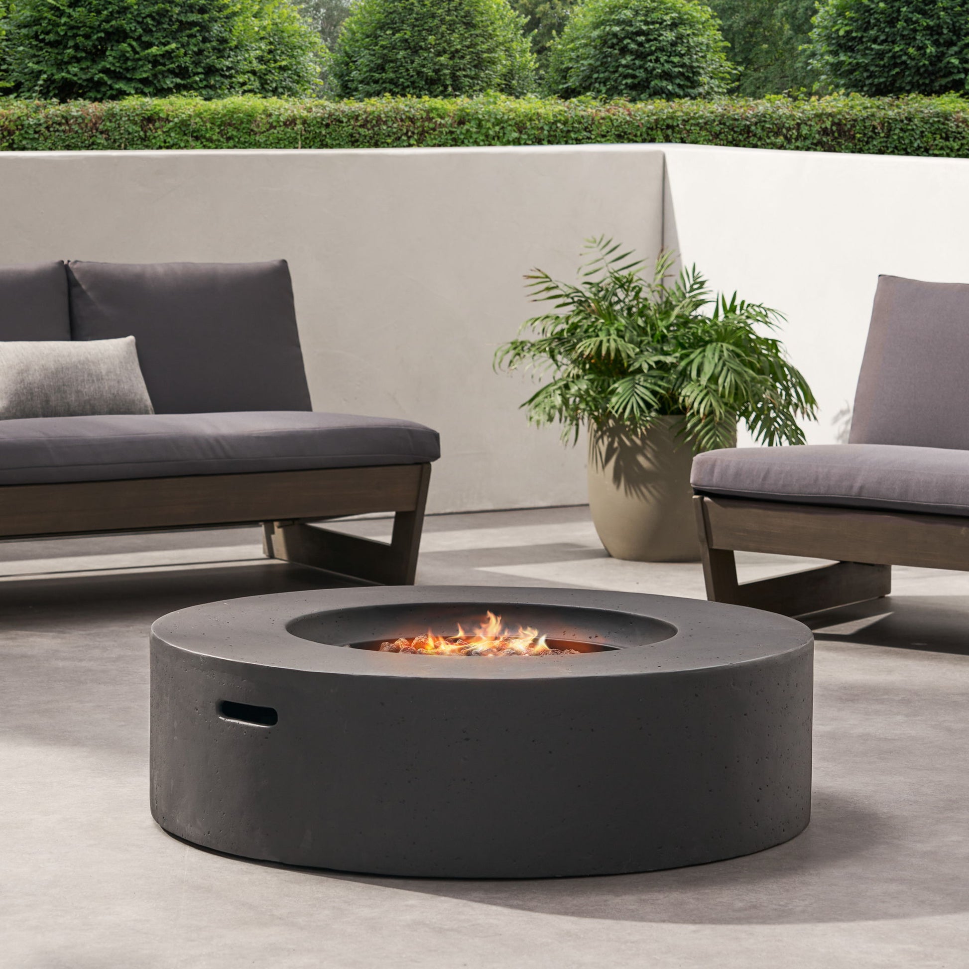 A modern patio with gray seating surrounds the Modern Lightweight Concrete Outdoor Circular Fire Pit 50,000 Btu, accented by a potted plant, lush greenery, and a white wall backdrop.