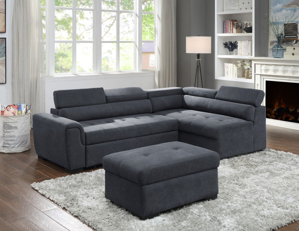 A modern living room featuring the Haris Fabric Sleeper Sofa Sectional with Adjustable Headrest and Storage Ottoman in dark gray on a white shag rug, wooden floor, large windows with curtains, lit floor lamp, bookshelves, and a fireplace.