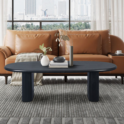 A modern living room with a brown leather sofa, plaid blanket, and a Tapered Tabletop Coffee Table in Antique Black featuring vases and a book. Large windows reveal a cityscape view in the background.