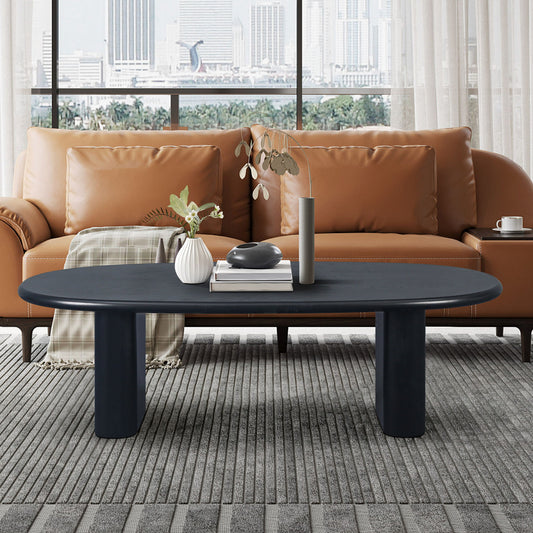 A modern living room with a brown leather sofa, plaid blanket, and a Tapered Tabletop Coffee Table in Antique Black featuring vases and a book. Large windows reveal a cityscape view in the background.