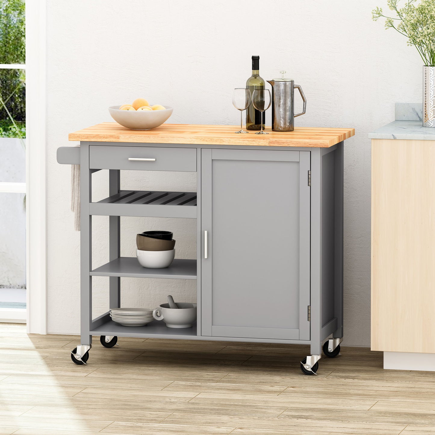 The Contemporary Design Kitchen Cart With Wheels features a gray finish, wooden top, open shelves, and a sliding door. On the counter are glasses, a carafe, and lemons, while bowls and plates are stored on the lower shelves.