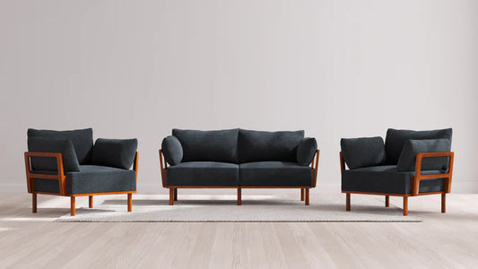 The Indoor Jack 2-Seater Sofa + Armchair Set in Balsa features a sleek and modern design with a dark gray finish. It includes a sofa and two matching armchairs with elegant wooden frames, all arranged on a light-colored rug atop a wooden floor, set against a plain, light gray wall.
