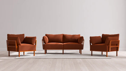 The Indoor Jack 2-Seater Sofa + Armchair Set in Balsa, with its rust-colored upholstery and wooden frames, is elegantly arranged on a light gray area rug. The setting features a plain, light-colored wall and a wooden floor in the background.