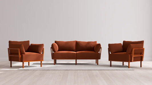 The Indoor Jack 2-Seater Sofa + Armchair Set in Balsa, with its rust-colored upholstery and wooden frames, is elegantly arranged on a light gray area rug. The setting features a plain, light-colored wall and a wooden floor in the background.