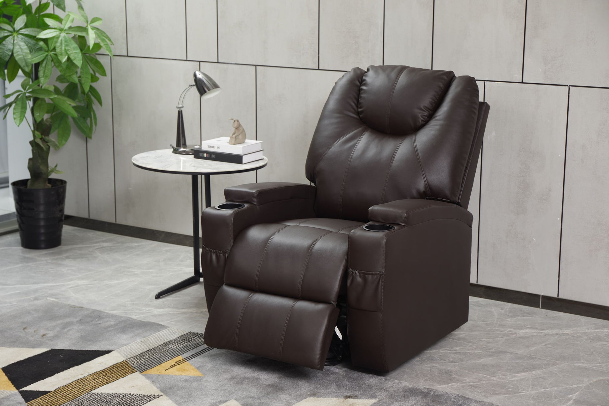 A Modern Recliner Chair With Cup Holders and remote control sits next to a small round side table with books, a lamp, and décor. Nearby, a green potted plant adds freshness to the stylish, tiled room.
