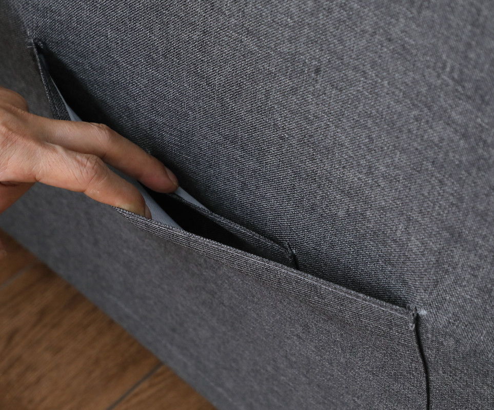A hand places a smartphone into the side pocket of the Zoey Convertible Sleeper Chair, which sits on a wooden floor.