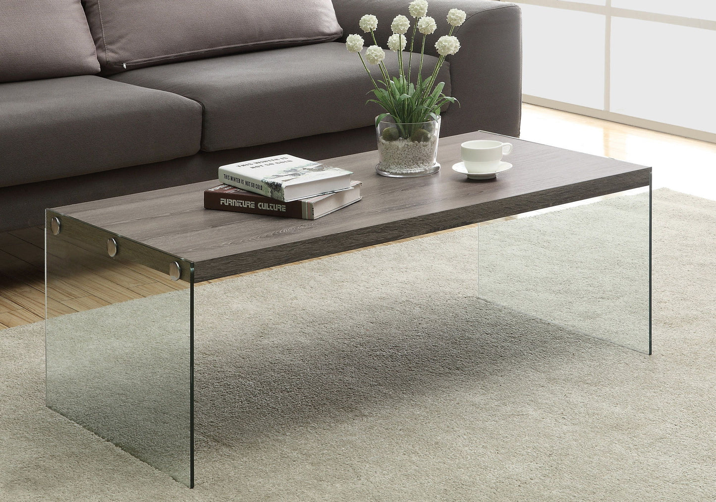 A Coffee Table Rectangular For Living Room Clear Tempered Glass with a wooden top stands on a beige rug, holding books, a white cup and saucer, and a glass vase with white flowers. A gray sofa is in the background.