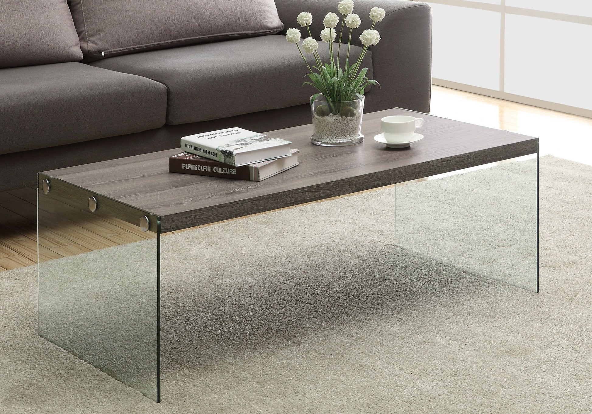 A Coffee Table Rectangular For Living Room Clear Tempered Glass with a wooden top stands on a beige rug, holding books, a white cup and saucer, and a glass vase with white flowers. A gray sofa is in the background.