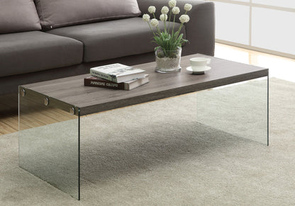 A Coffee Table Rectangular For Living Room Clear Tempered Glass with a wooden top stands on a beige rug, holding books, a white cup and saucer, and a glass vase with white flowers. A gray sofa is in the background.