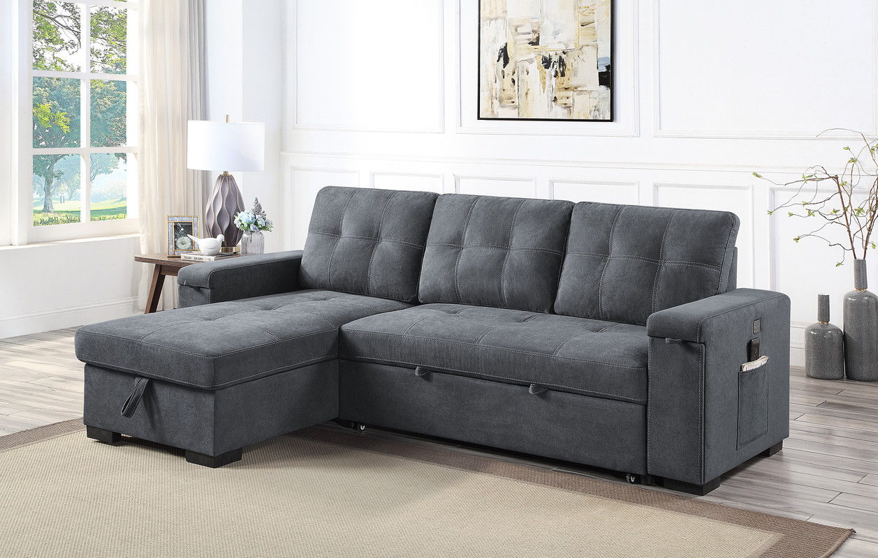 A modern living room featuring the Toby Woven Fabric Reversible Sleeper Sectional Sofa with tufted cushions, storage chaise, cup holder, charging ports, and pockets; styled with a beige rug, side table and lamp, plant, white-curtained window, and abstract wall art.
