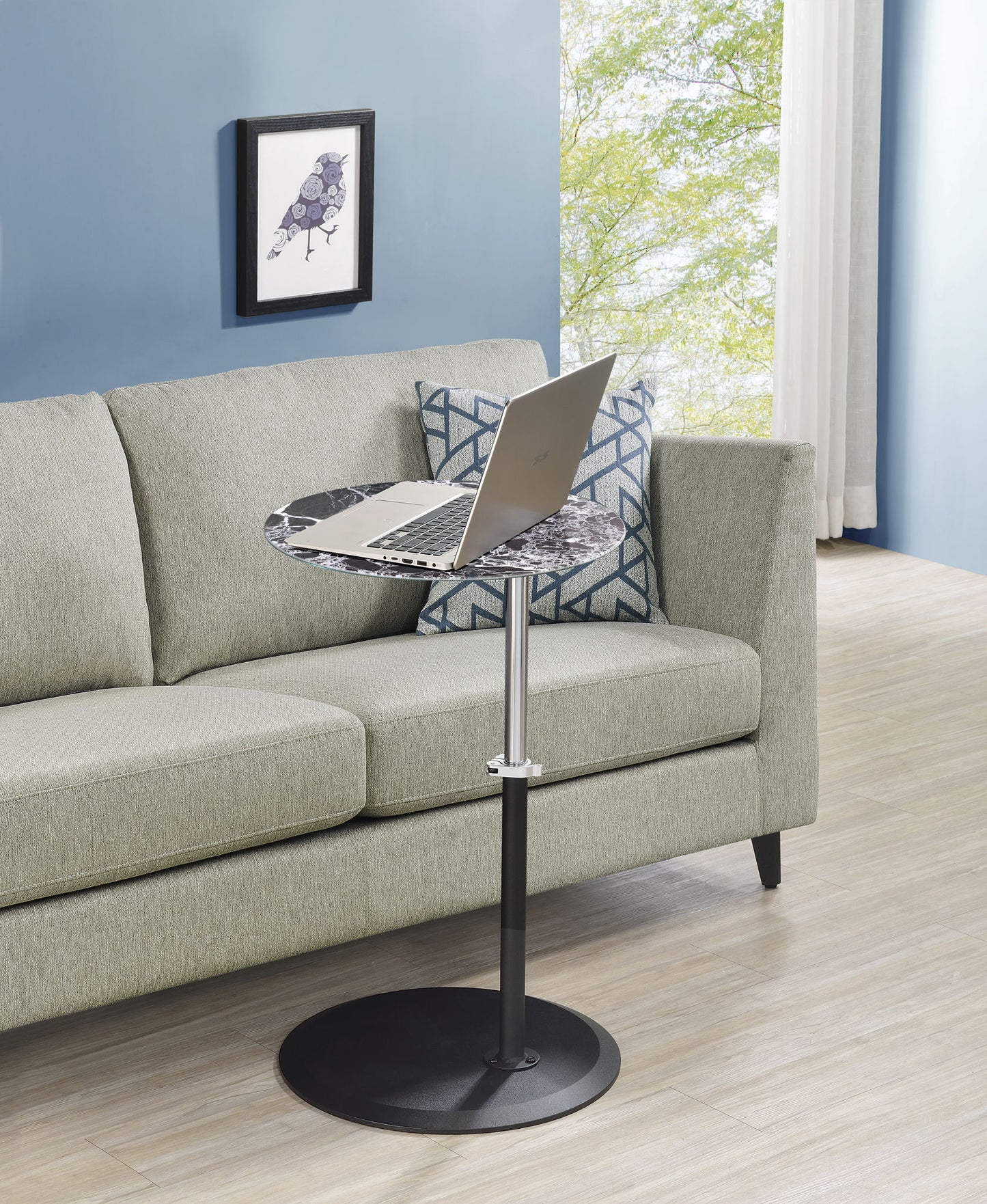 The Orbit 15.5" End Table with a height-adjustable marble-textured top and laptop sits before a light gray sofa with patterned pillows. A framed bird print hangs on the blue wall, and a window reveals greenery outside.