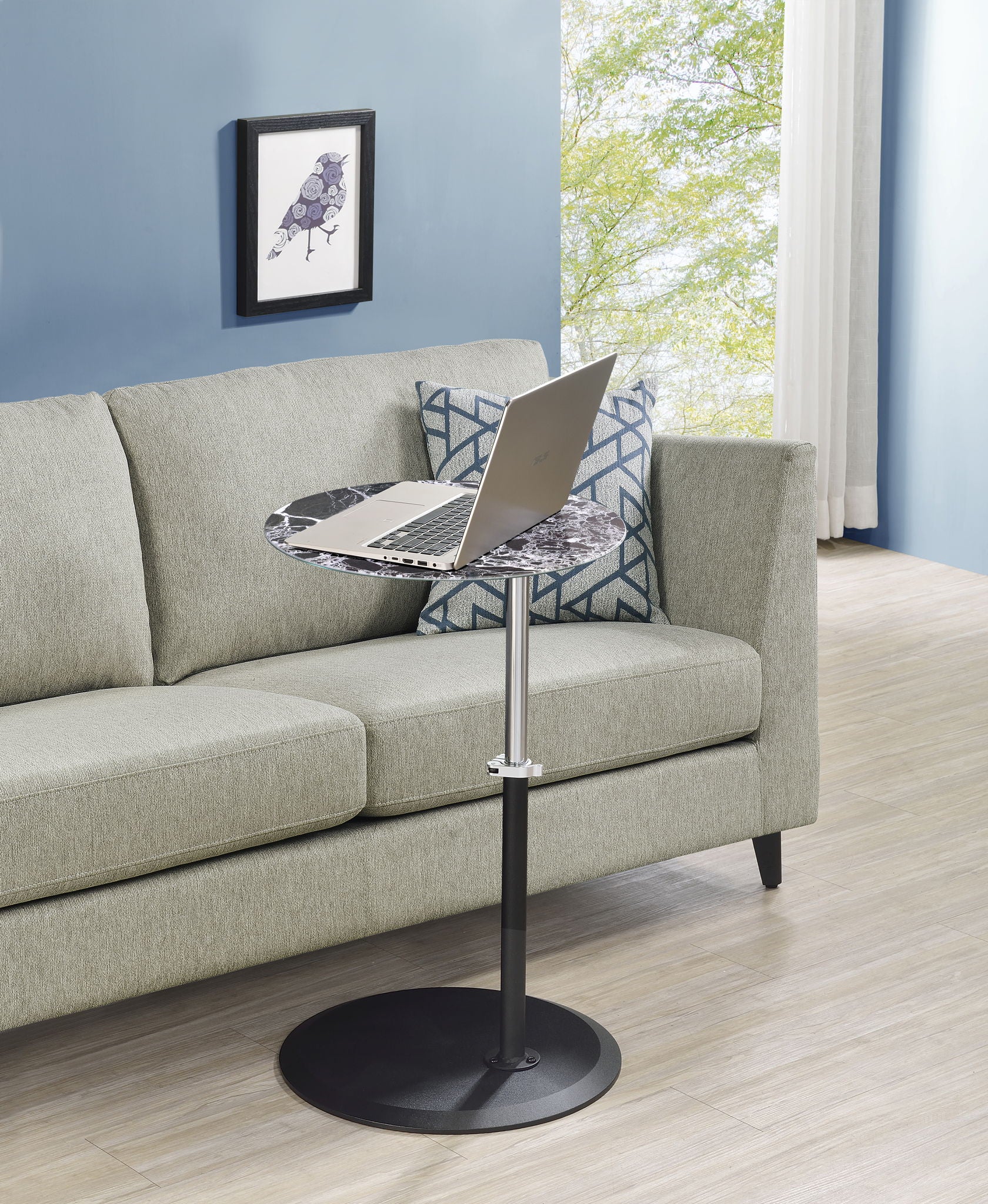 The Orbit 15.5" End Table with a height-adjustable marble-textured top and laptop sits before a light gray sofa with patterned pillows. A framed bird print hangs on the blue wall, and a window reveals greenery outside.