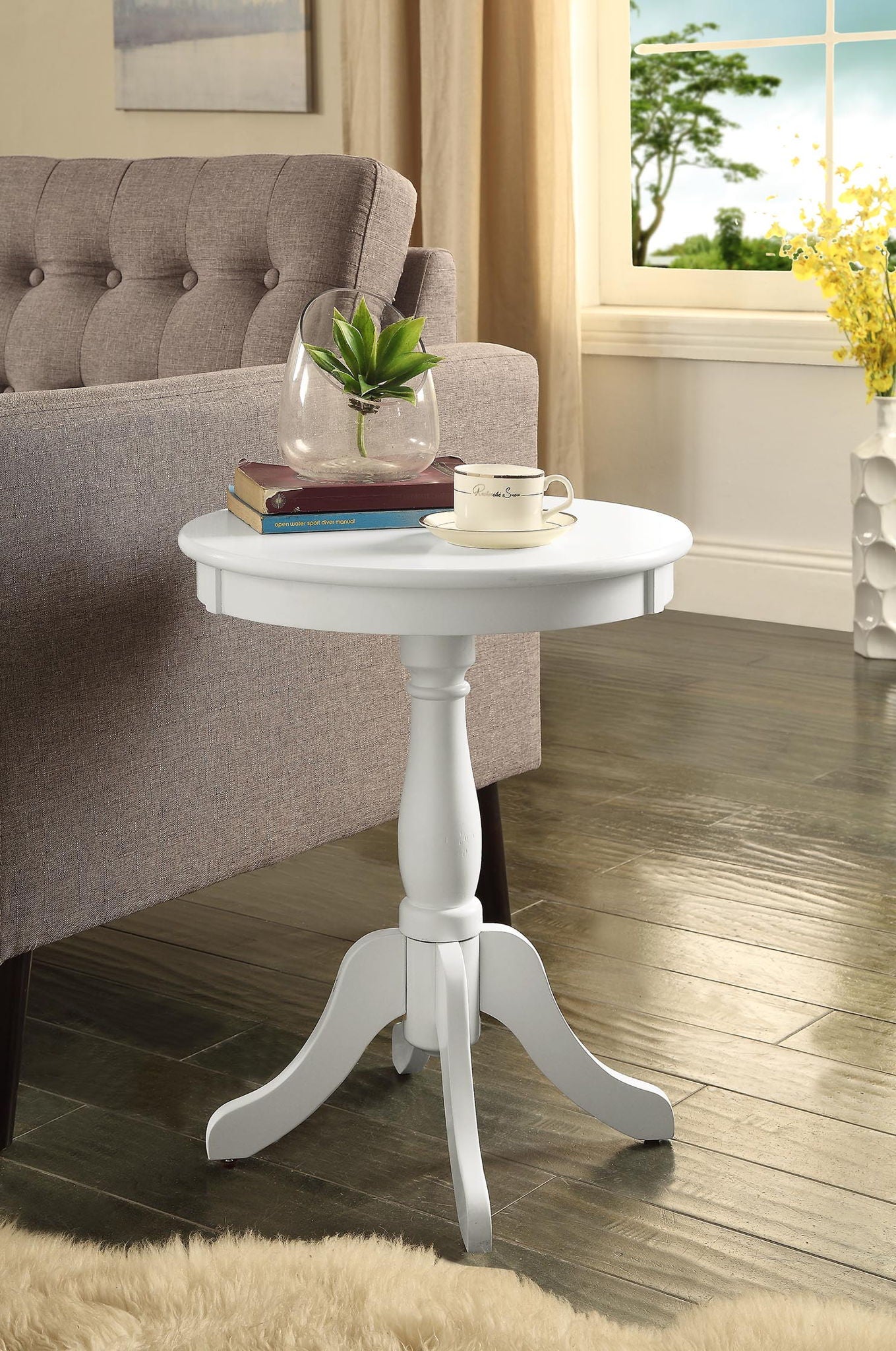 The Alger Accent Table in white sits by a gray sofa, holding a glass vase with a plant, a book, and a teacup. Behind it, wooden flooring leads to a window with curtains and yellow flowers in the background.