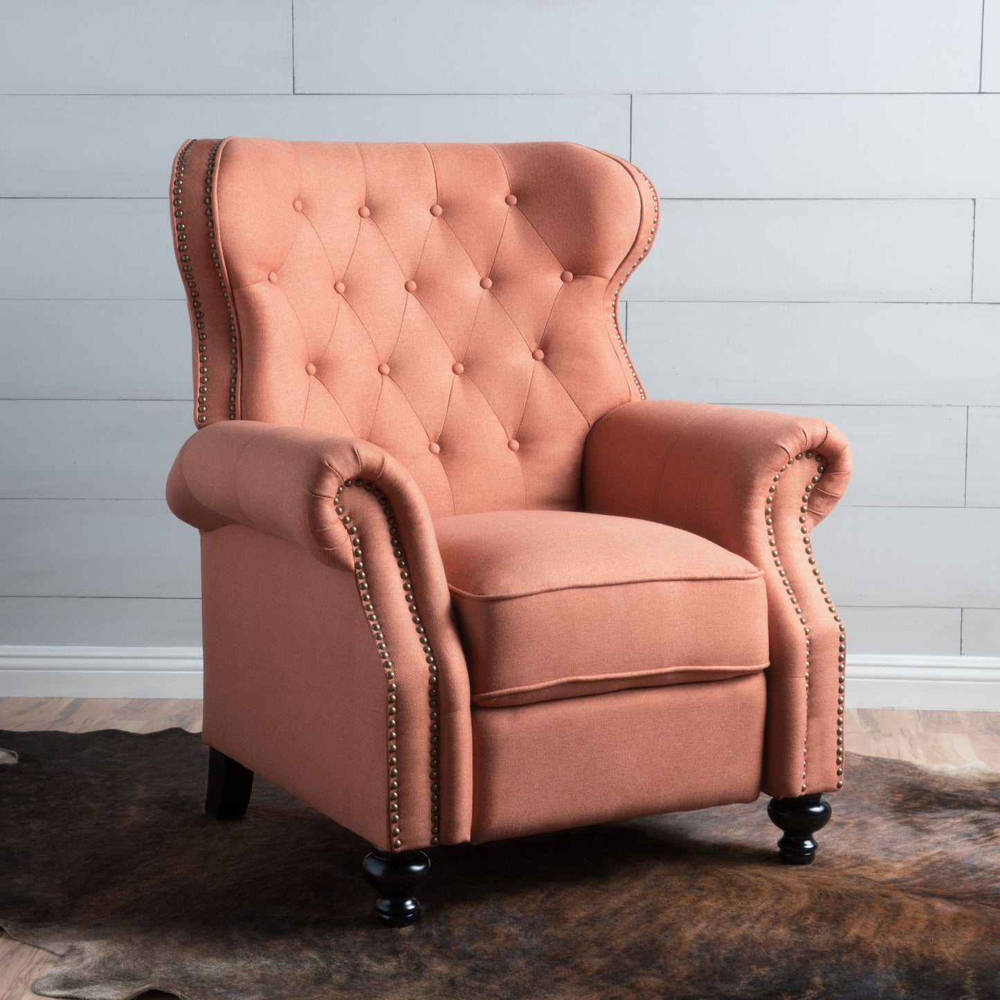 A salmon pink Contemporary Style Push Back Recliner with button tufted stitching and nailhead trim stands on dark wooden legs atop a cowhide rug on a wood floor, set against a white shiplap wall.