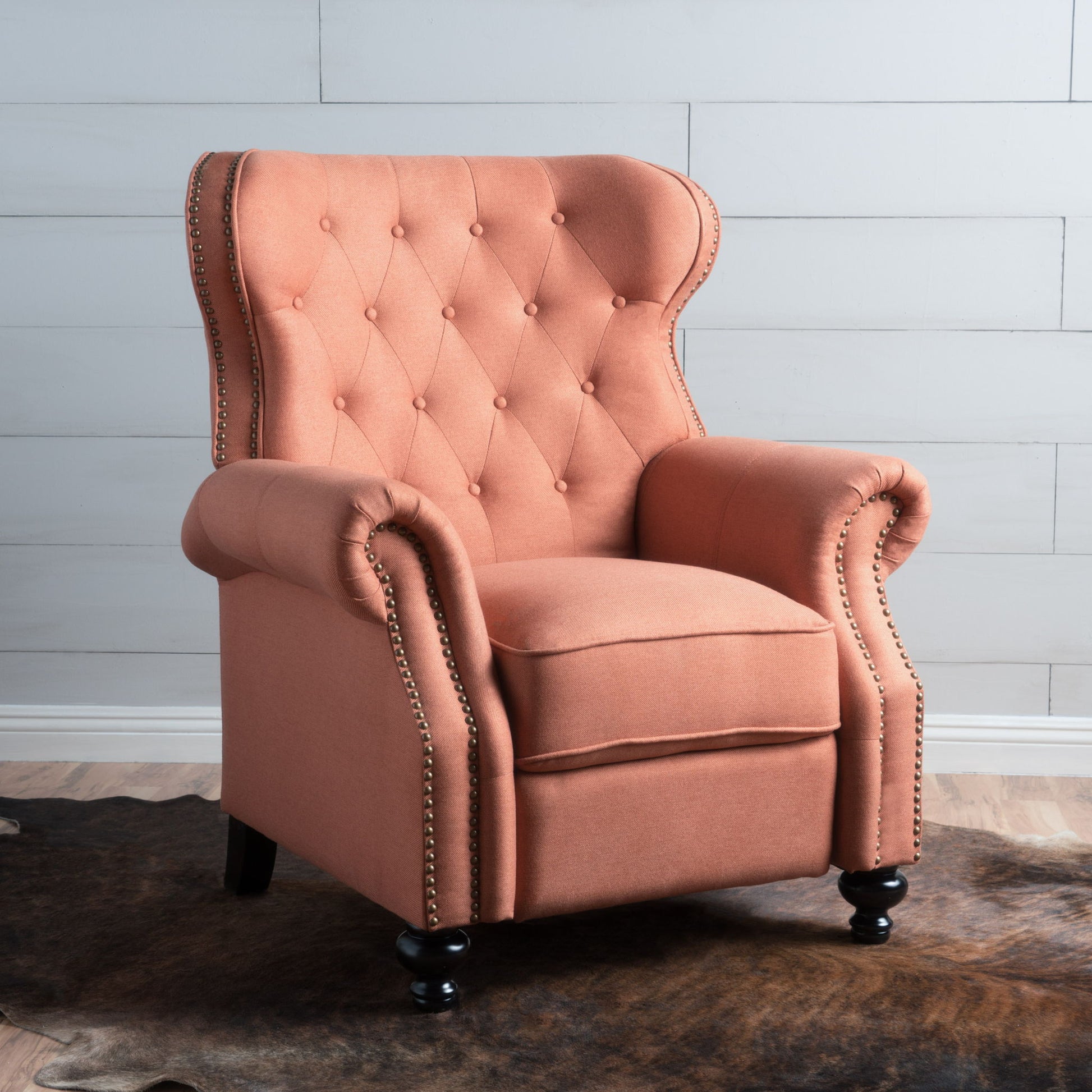 A salmon pink Contemporary Style Push Back Recliner with button tufted stitching and nailhead trim stands on dark wooden legs atop a cowhide rug on a wood floor, set against a white shiplap wall.