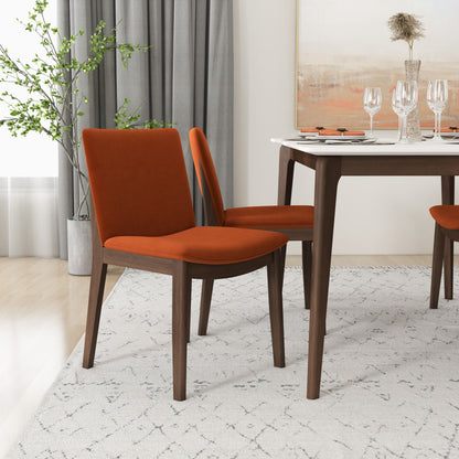 A dining room features a table set with wine glasses, plates, and cutlery. Nearby on a patterned rug are two Laura mid-century modern solid wood dining chairs with orange cushions. A plant sits in the corner, with gray curtains and a painting visible in the background.