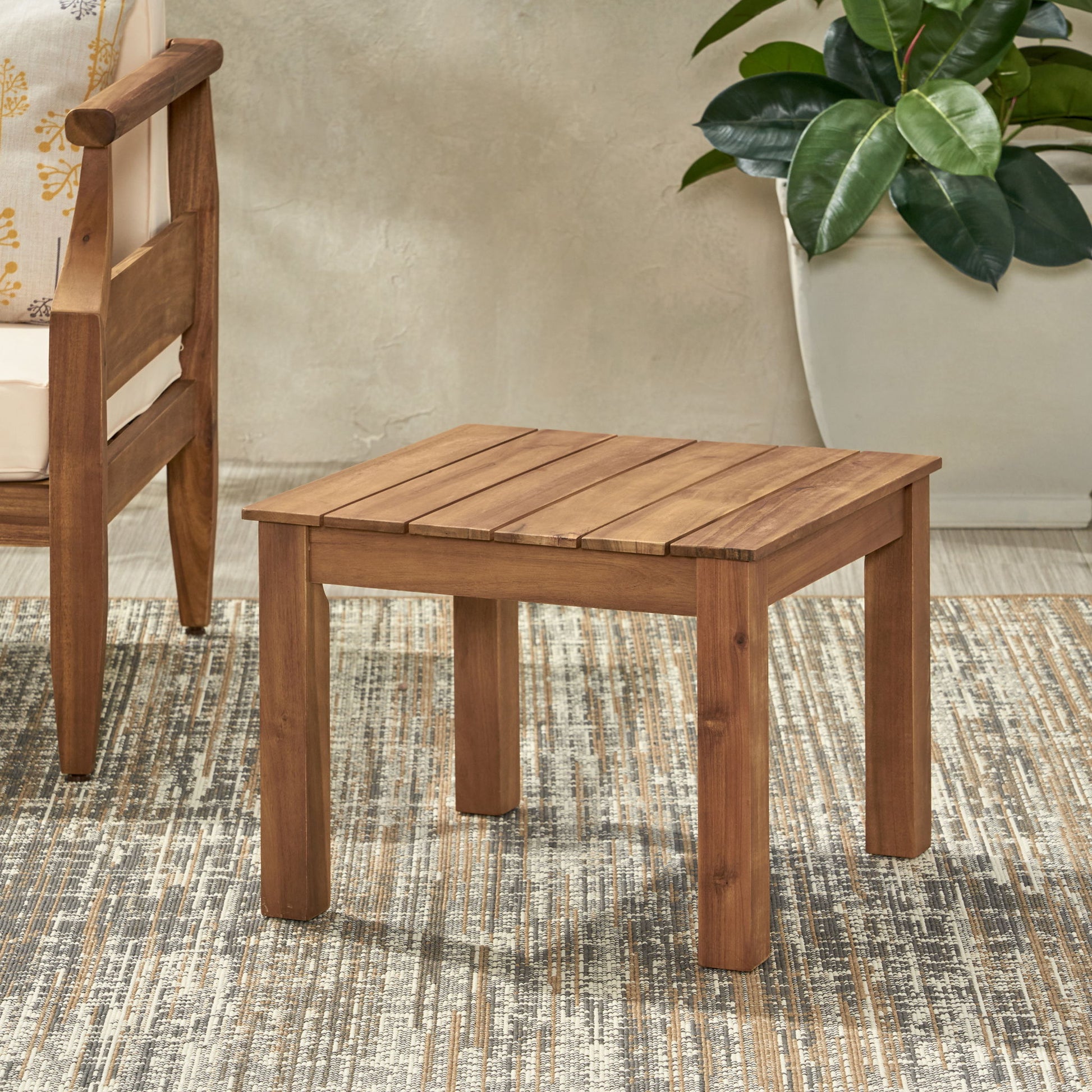 The Halcyon - Outdoor Side Table With Slat Design sits on a patterned rug next to a wooden chair and a large potted plant, set against a light-colored wall.