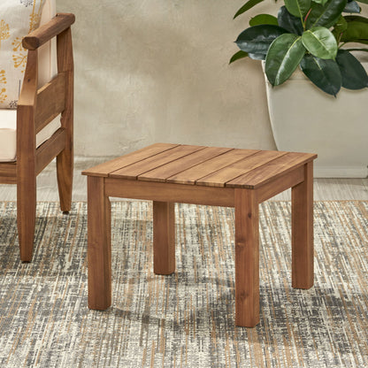 The Halcyon - Outdoor Side Table With Slat Design sits on a patterned rug next to a wooden chair and a large potted plant, set against a light-colored wall.