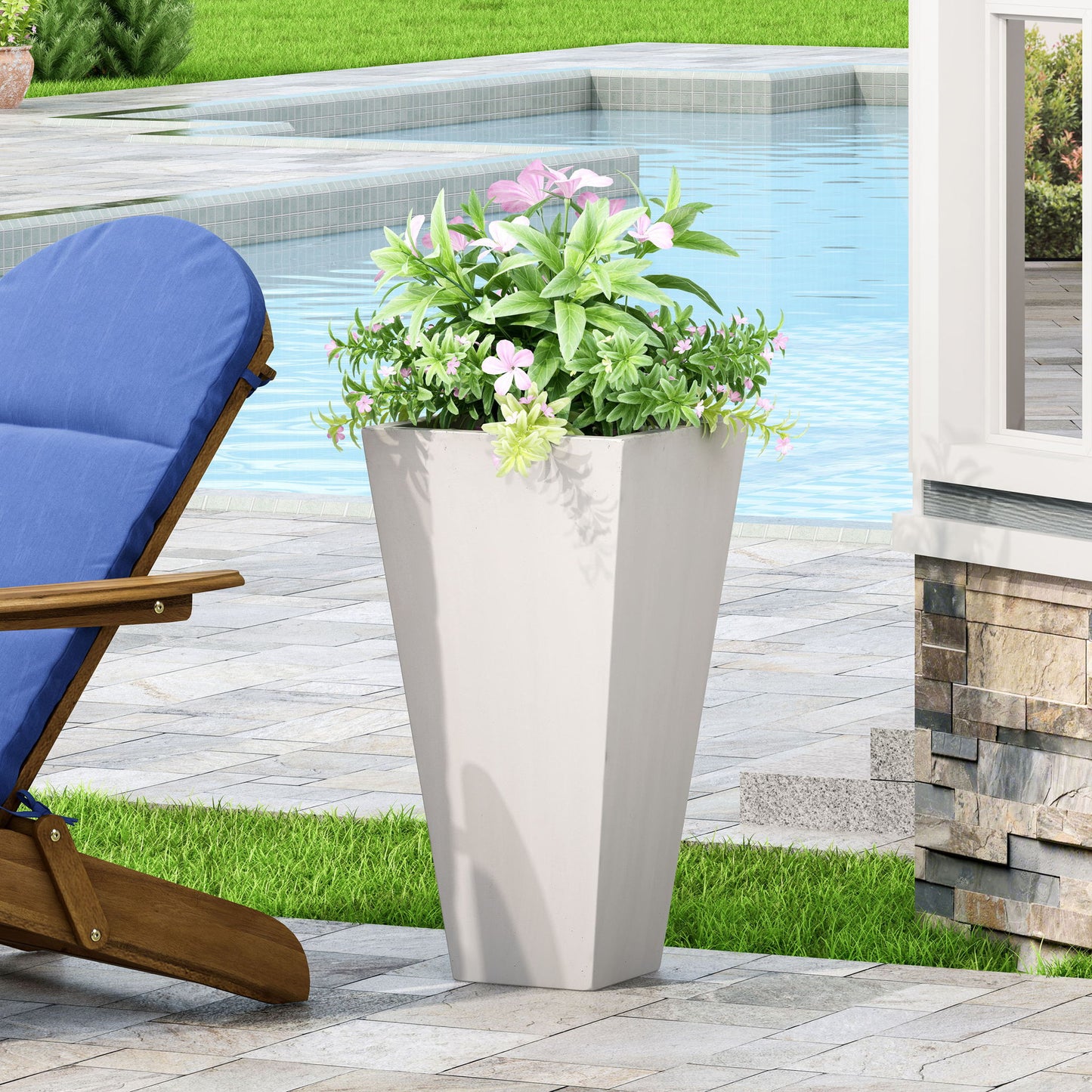 An Outdoor Modern Cast Stone Planter with green foliage and pink flowers sits on a stone patio beside a blue-cushioned wooden lounge chair, with a pool and lush greenery in the background.