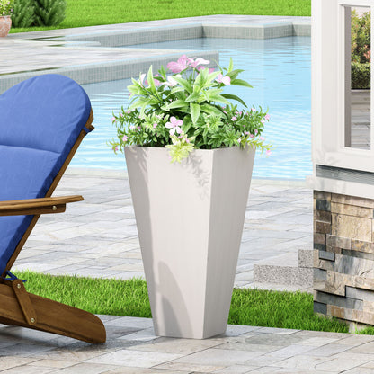 An Outdoor Modern Cast Stone Planter with green foliage and pink flowers sits on a stone patio beside a blue-cushioned wooden lounge chair, with a pool and lush greenery in the background.