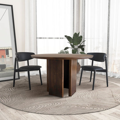 The Alden Round Dining Table in walnut is paired with two black chairs, set on a patterned rug in a bright room with large windows, white curtains, a potted plant, and a framed photo against the wall.