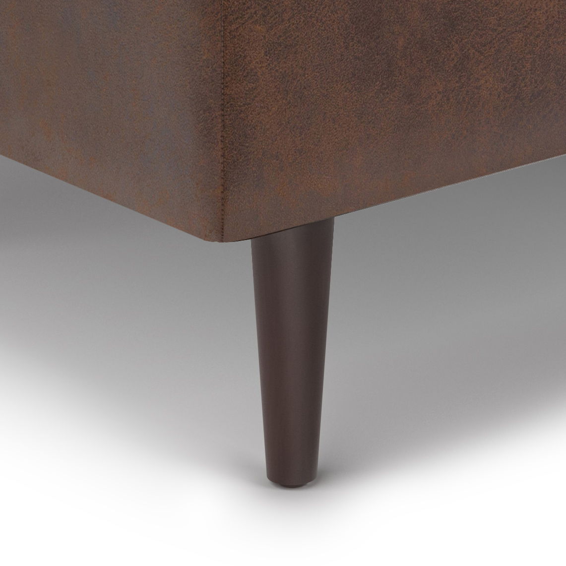 Close-up of the Shay - Large Square Coffee Table Storage Ottoman (Mid-Century Style) in brown upholstery, highlighting a single dark brown tapered wooden leg on a light grey floor.