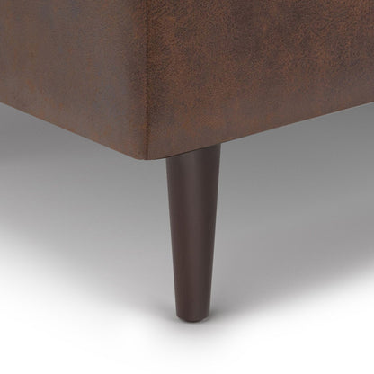 Close-up of the Shay - Large Square Coffee Table Storage Ottoman (Mid-Century Style) in brown upholstery, highlighting a single dark brown tapered wooden leg on a light grey floor.