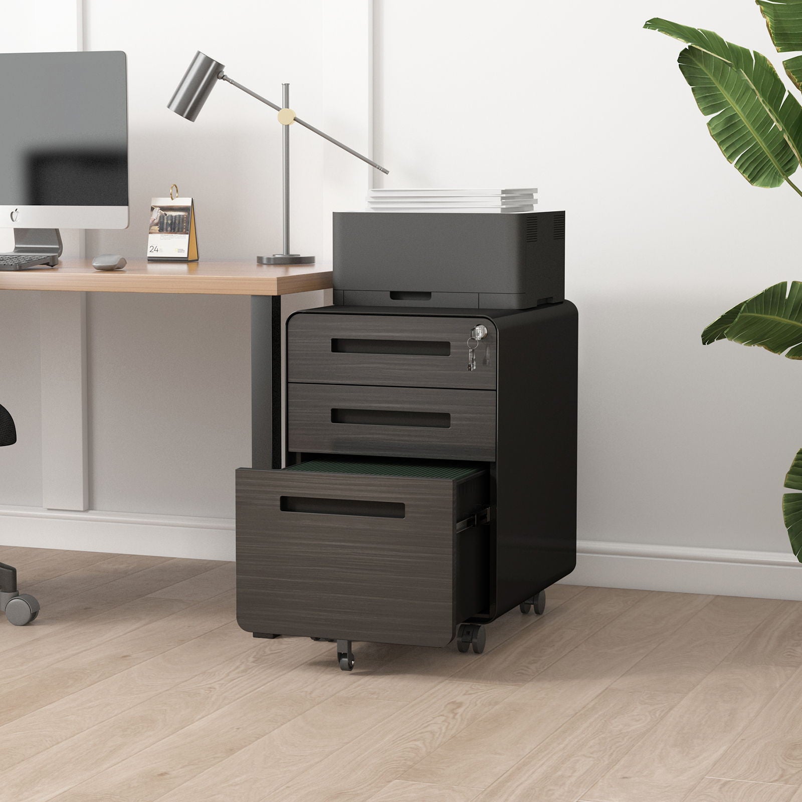 A modern office features the 3 Drawer Mobile File Cabinet Under Desk Office, a black, 5-wheel steel cabinet with three drawers—including one open with files—plus a printer and paper on top. A plant sits nearby for a stylish touch.