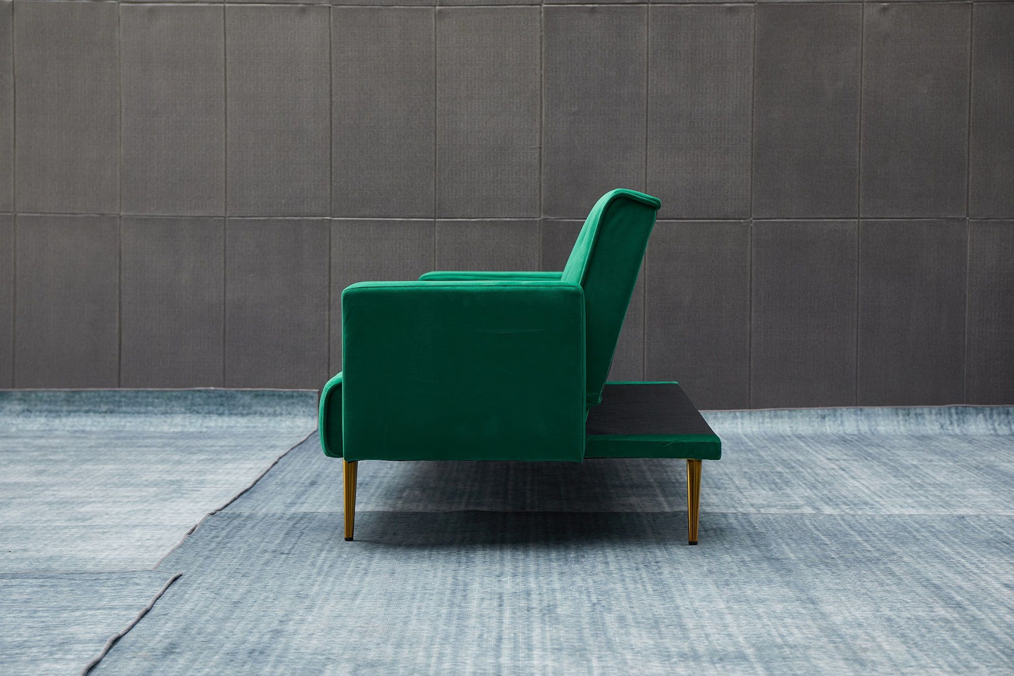The Comfort Compact Design Sofa Bed in green upholstery with gold legs is displayed in profile on a blue textured rug, set against a gray paneled wall.