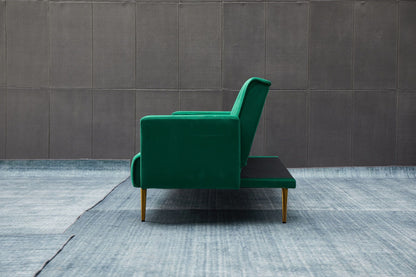 The Comfort Compact Design Sofa Bed in green upholstery with gold legs is displayed in profile on a blue textured rug, set against a gray paneled wall.