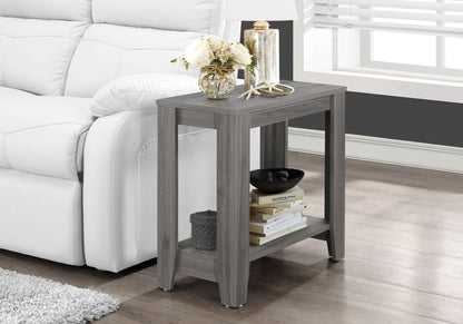An Accent Table, Side Transitional with an elegant design holds a flower vase, decorative candle, and lamp on top, with books, a bowl, and basket on the lower shelf. It sits beside a white sofa in a bright living room.