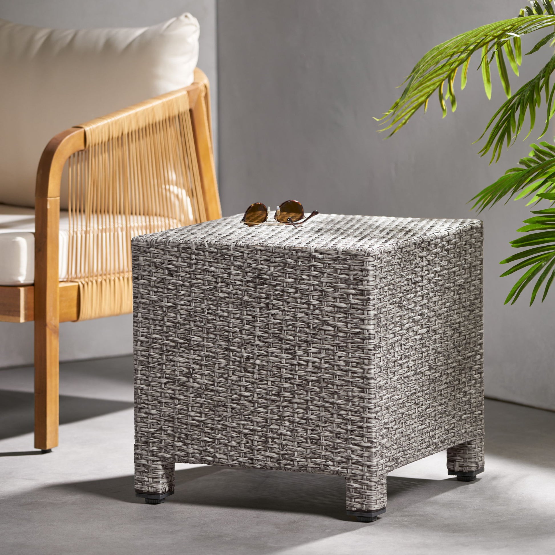 The Puerta Minimalist Style Accent Table, topped with sunglasses, sits beside a wooden chair with cream cushions and a green leafy plant in a sunlit room.