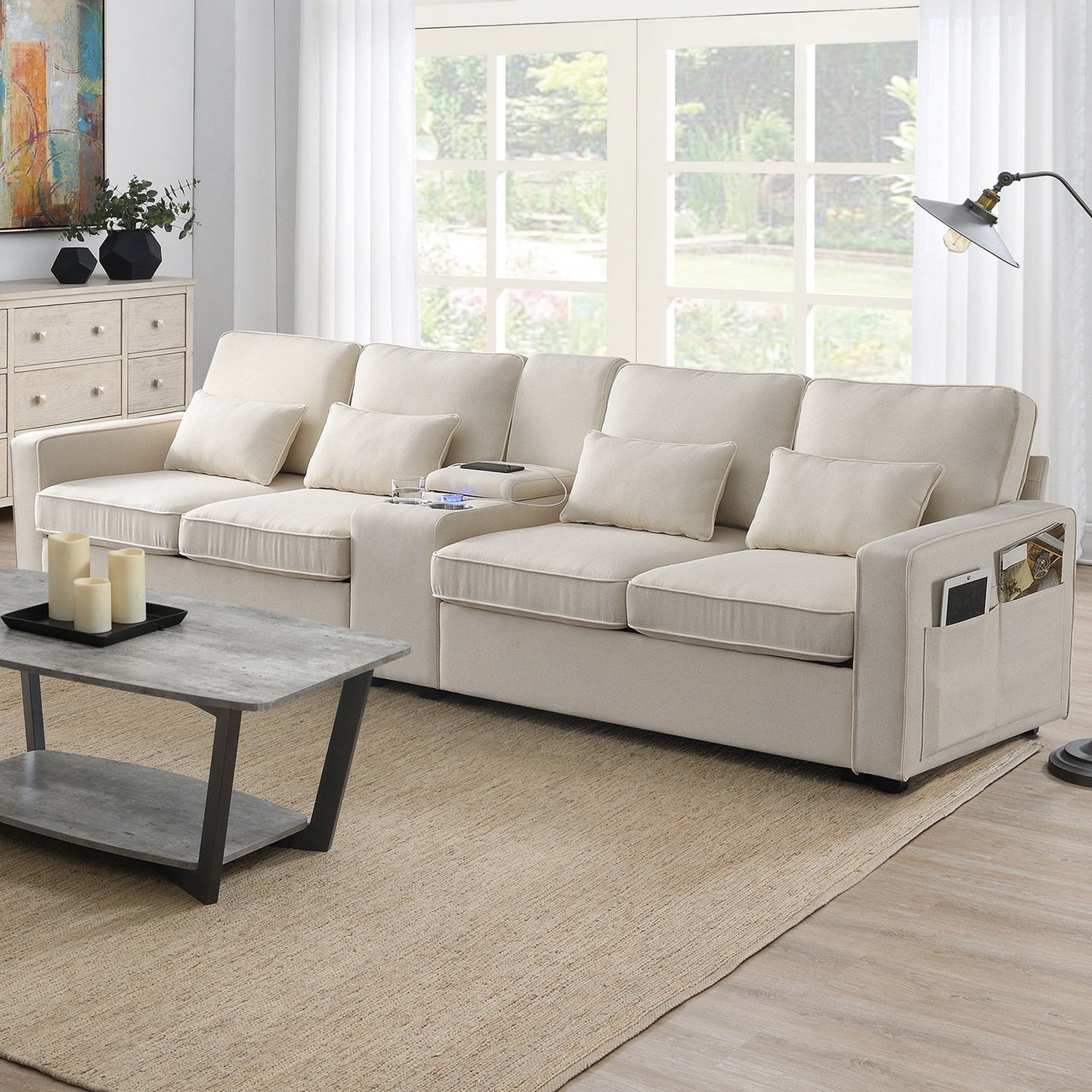 A modern living room features the Upholstered Sofa With Console—an elegant 4-seat linen couch with 4 pillows, built-in cupholders, USB charging ports (wired/wireless), and a cozy setup highlighted by a coffee table on a tan rug near large windows.