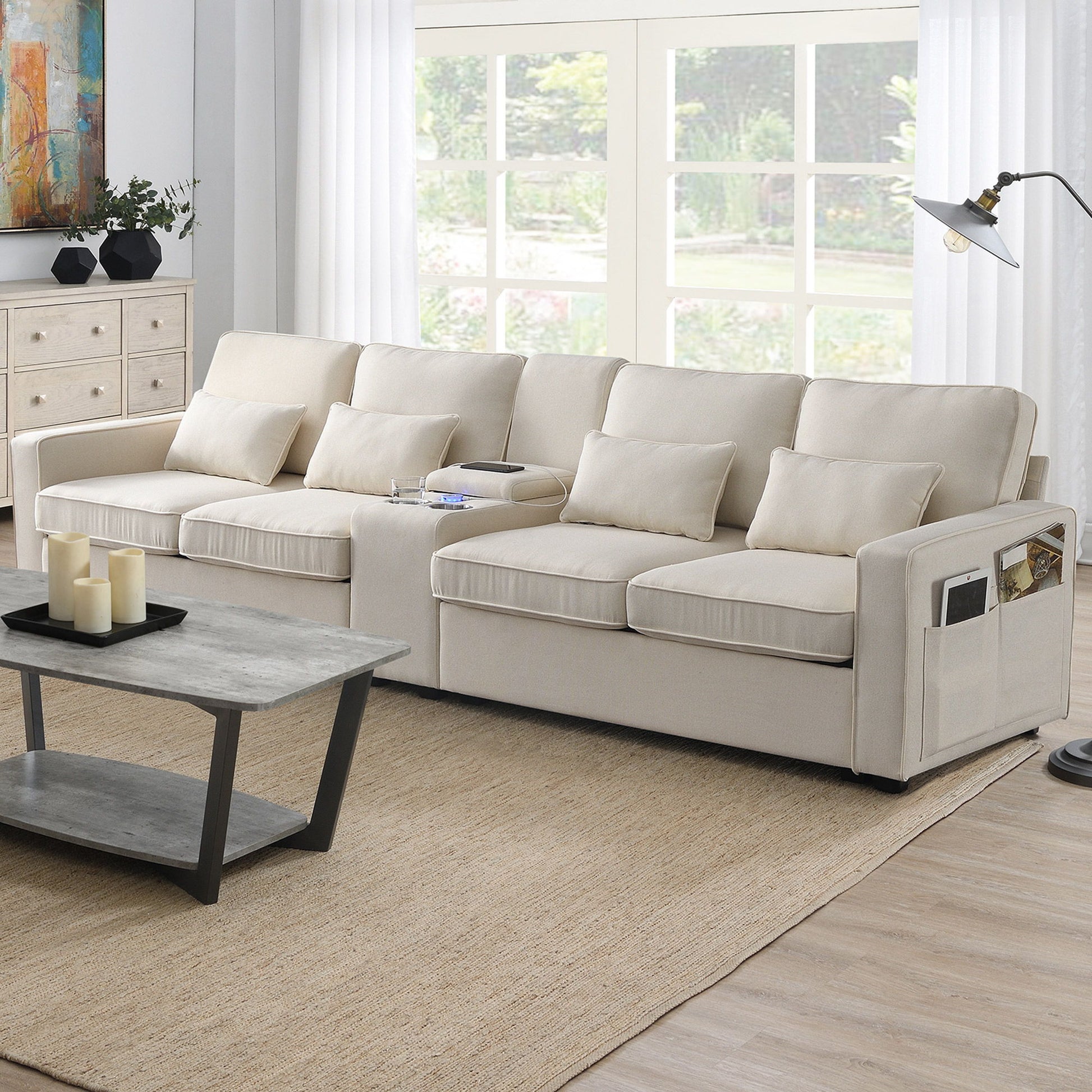 A modern living room features the Upholstered Sofa With Console—an elegant 4-seat linen couch with 4 pillows, built-in cupholders, USB charging ports (wired/wireless), and a cozy setup highlighted by a coffee table on a tan rug near large windows.