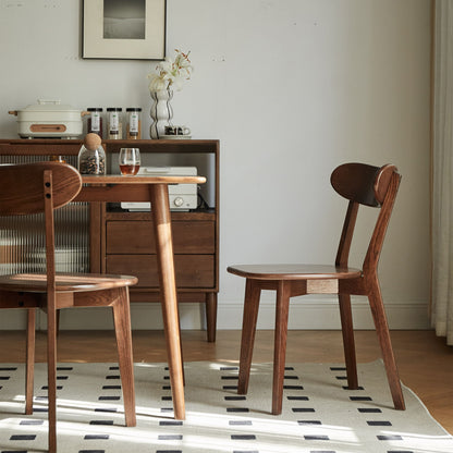 A cozy dining area with a Simple Dining Chair and solid table, a sideboard holding kitchen items, a framed photo on the wall, and a light rug with black rectangles over wooden floors. Sunlight streams in from the right.