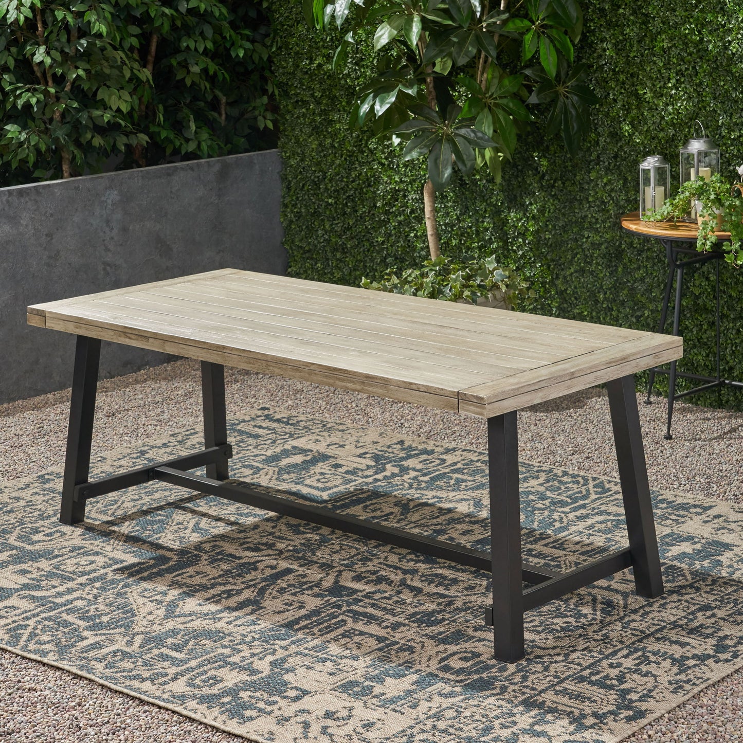 The Equilibrium Outdoor Dining Table with wooden frame and slat top sits on a patterned outdoor rug in the garden, surrounded by green plants and a small round table adorned with lanterns.