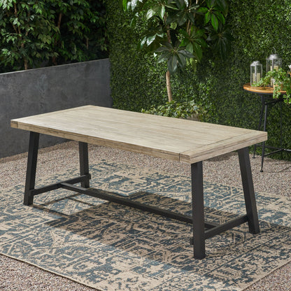 The Equilibrium Outdoor Dining Table with wooden frame and slat top sits on a patterned outdoor rug in the garden, surrounded by green plants and a small round table adorned with lanterns.