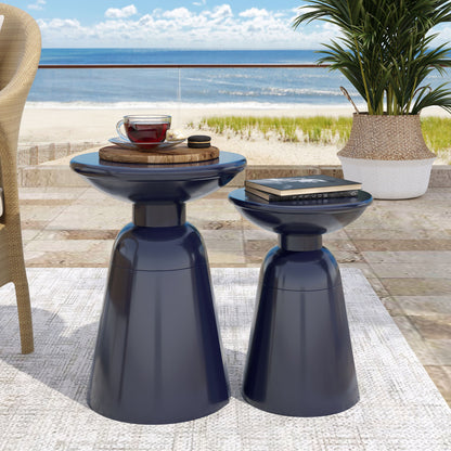 Two Serenita Outdoor Side Tables with a flared design (set of 2) in dark blue sit on a patio, topped with a tray, tea, and a book. Nearby are a potted plant and wicker chair, with an ocean view and blue sky in the background.