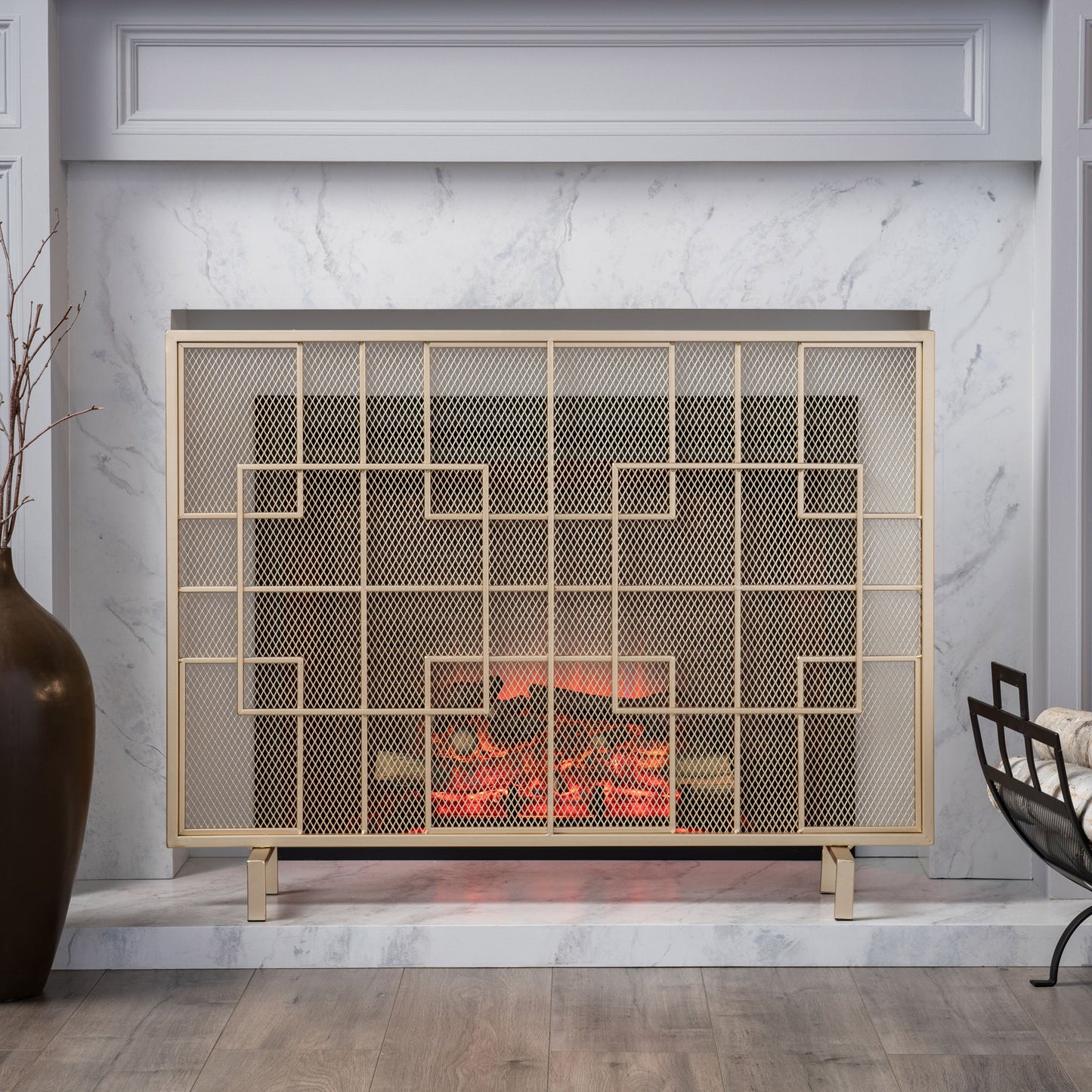 The Dorothy Modern Single Panel Iron Fire Screen features a sleek geometric design in gold, set within a white marble fireplace. A large dark vase with branches sits on the left, while a cozy chair draped with a blanket rests on the right.