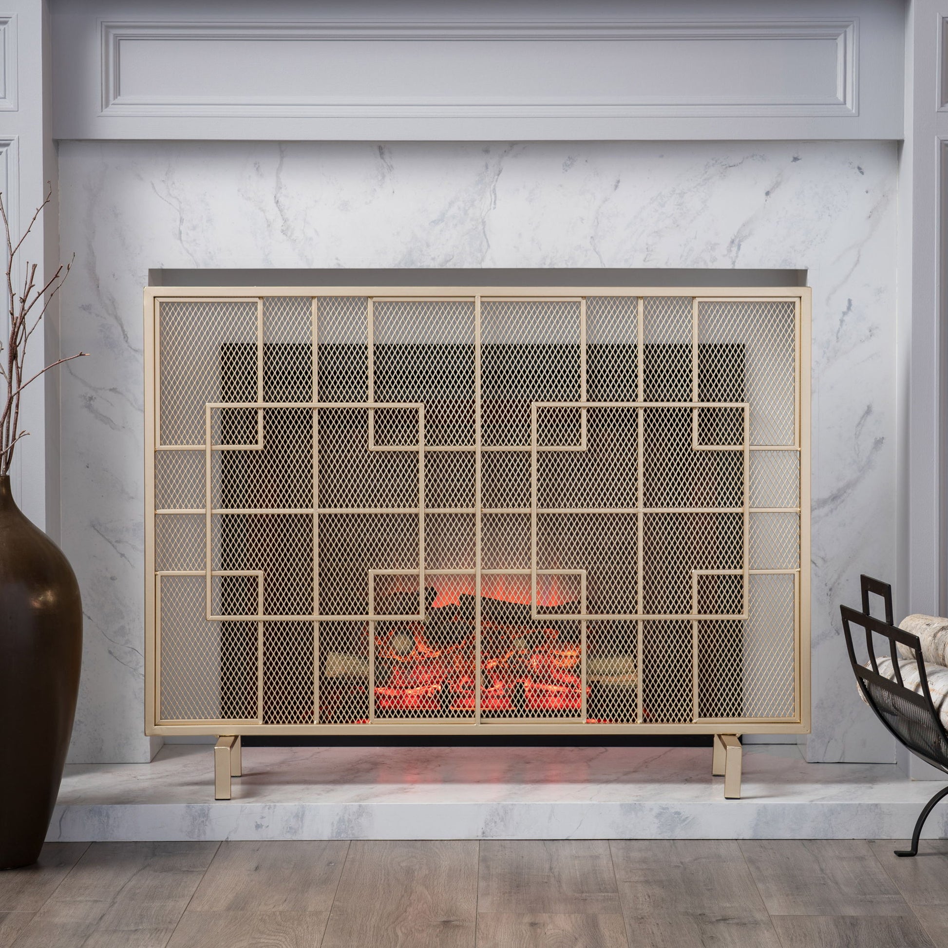 The Dorothy Modern Single Panel Iron Fire Screen features a sleek geometric design in gold, set within a white marble fireplace. A large dark vase with branches sits on the left, while a cozy chair draped with a blanket rests on the right.