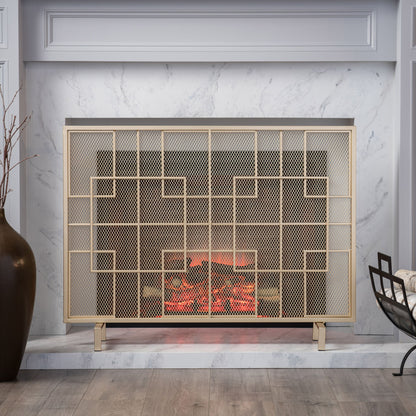 The Dorothy Modern Single Panel Iron Fire Screen features a sleek geometric design in gold, set within a white marble fireplace. A large dark vase with branches sits on the left, while a cozy chair draped with a blanket rests on the right.