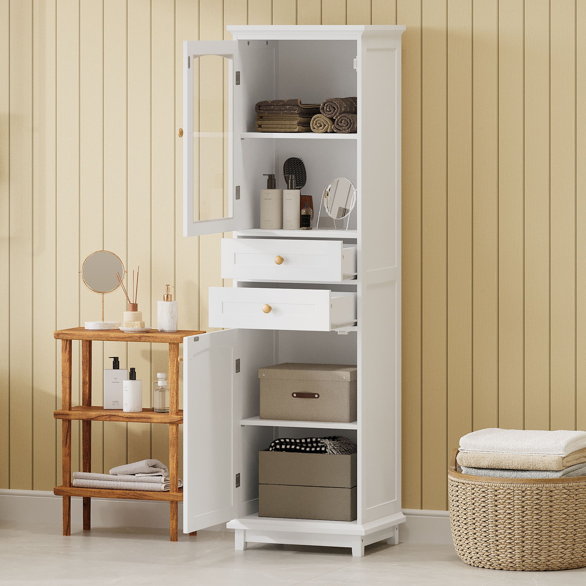 A tall white free-standing bathroom storage cabinet with glass doors, two drawers, and adjustable shelves displays towels and toiletries against a beige paneled wall. Wicker baskets sit on the floor beside it for extra storage.