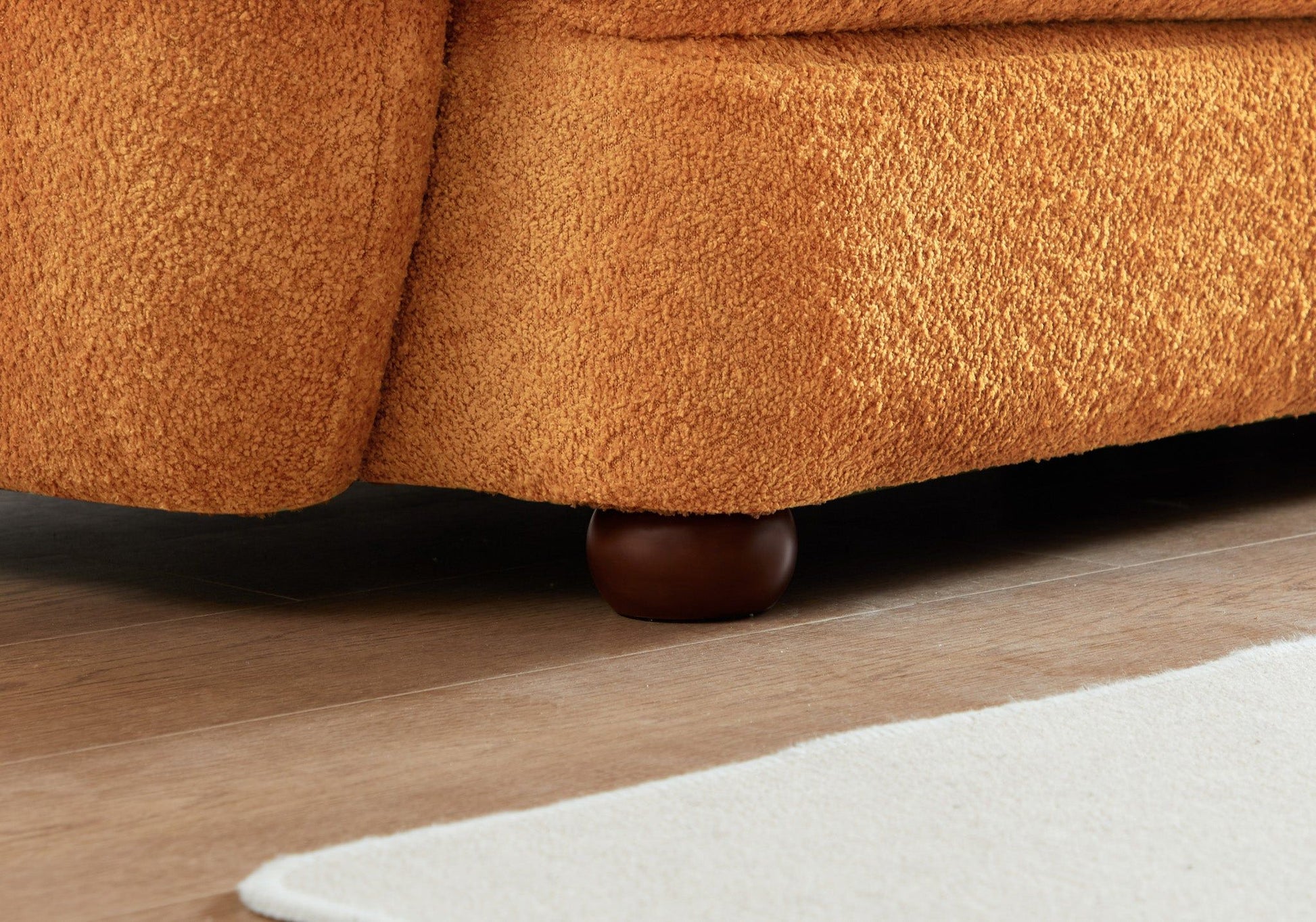 Close-up of the bottom corner of the Paton - Comfortable Sofa in orange texture, featuring a rounded dark wooden leg on a light wood floor beside a white rug.