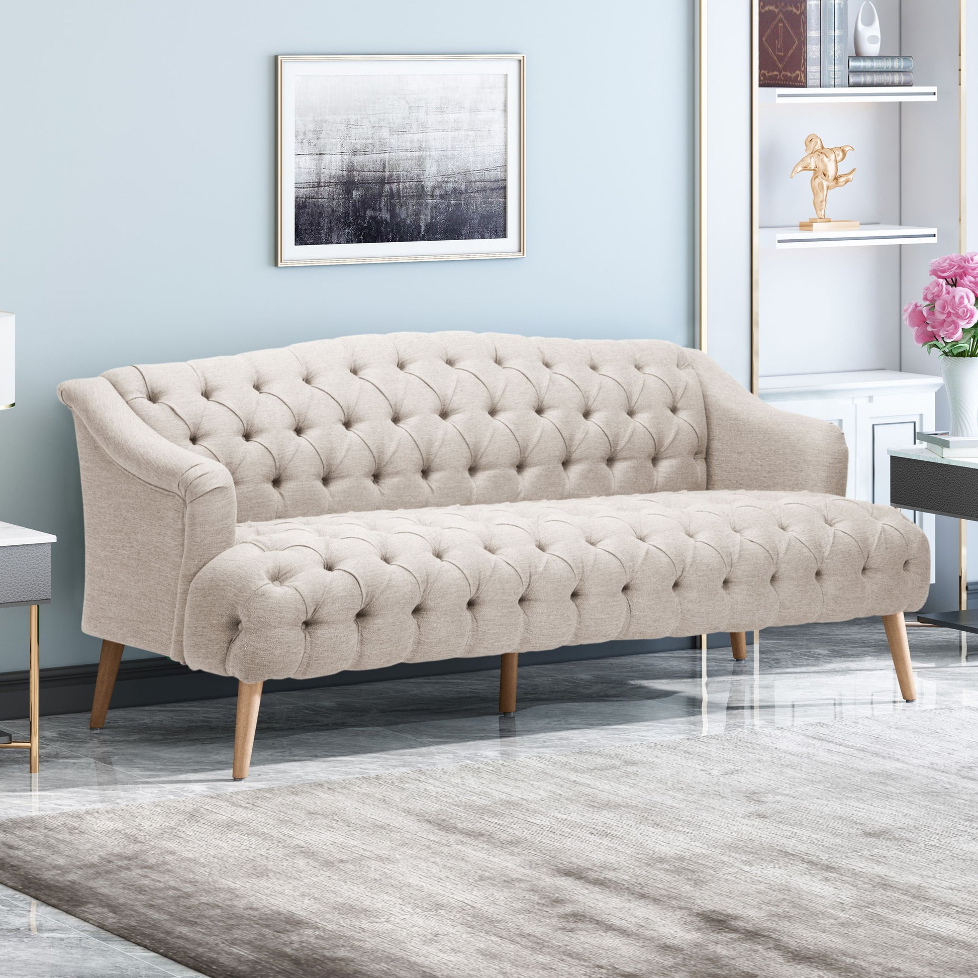 A modern living room featuring the Comfy 3 Seat Sofa with wooden legs, a gray rug, abstract wall art, shelves with books and décor, and a side table with a vase of pink flowers.