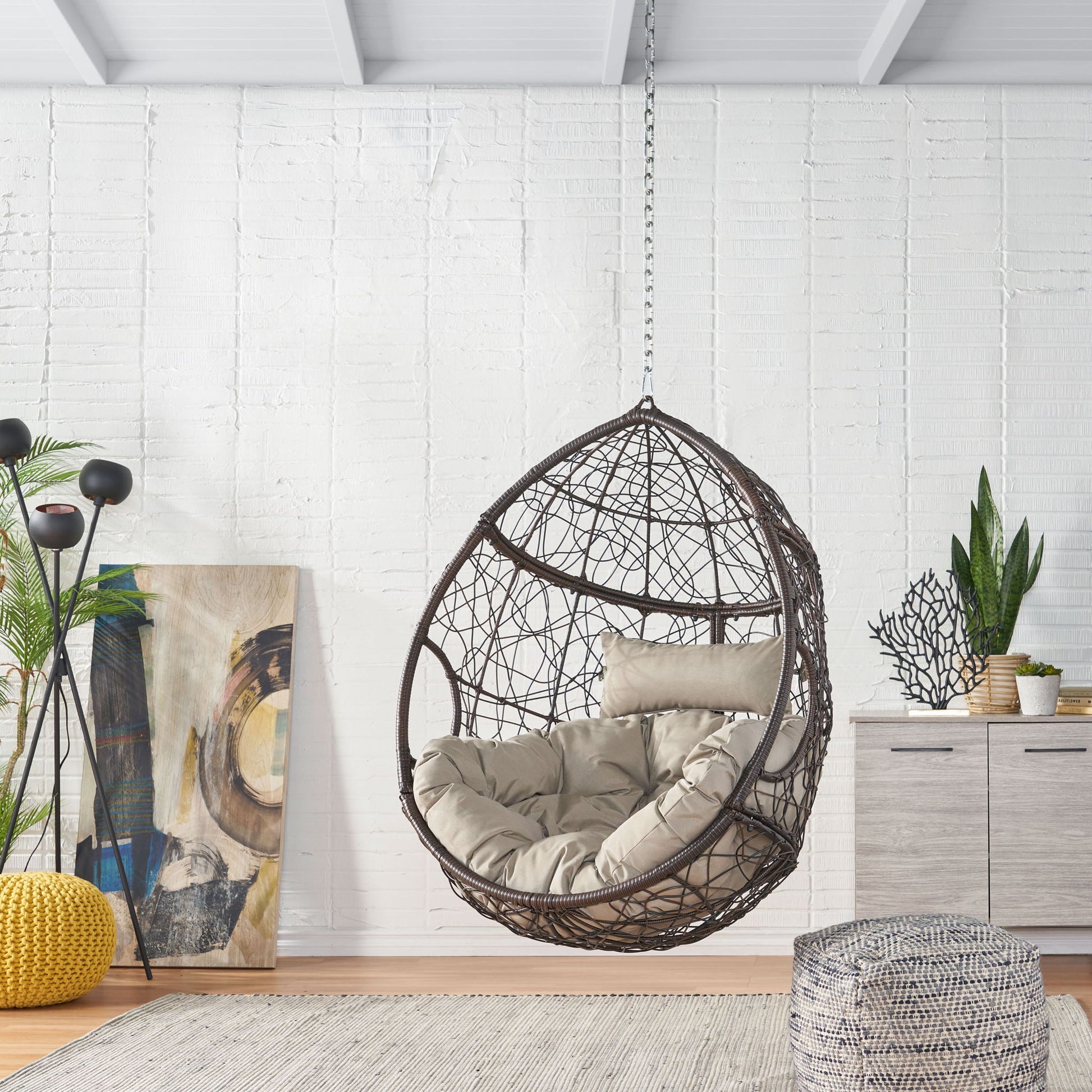 The Teardrop - Hanging Chair With 8Ft Chain, featuring beige cushions, hangs from the ceiling in a modern, light-filled room with white brick walls, artwork, plants, and contemporary decor.