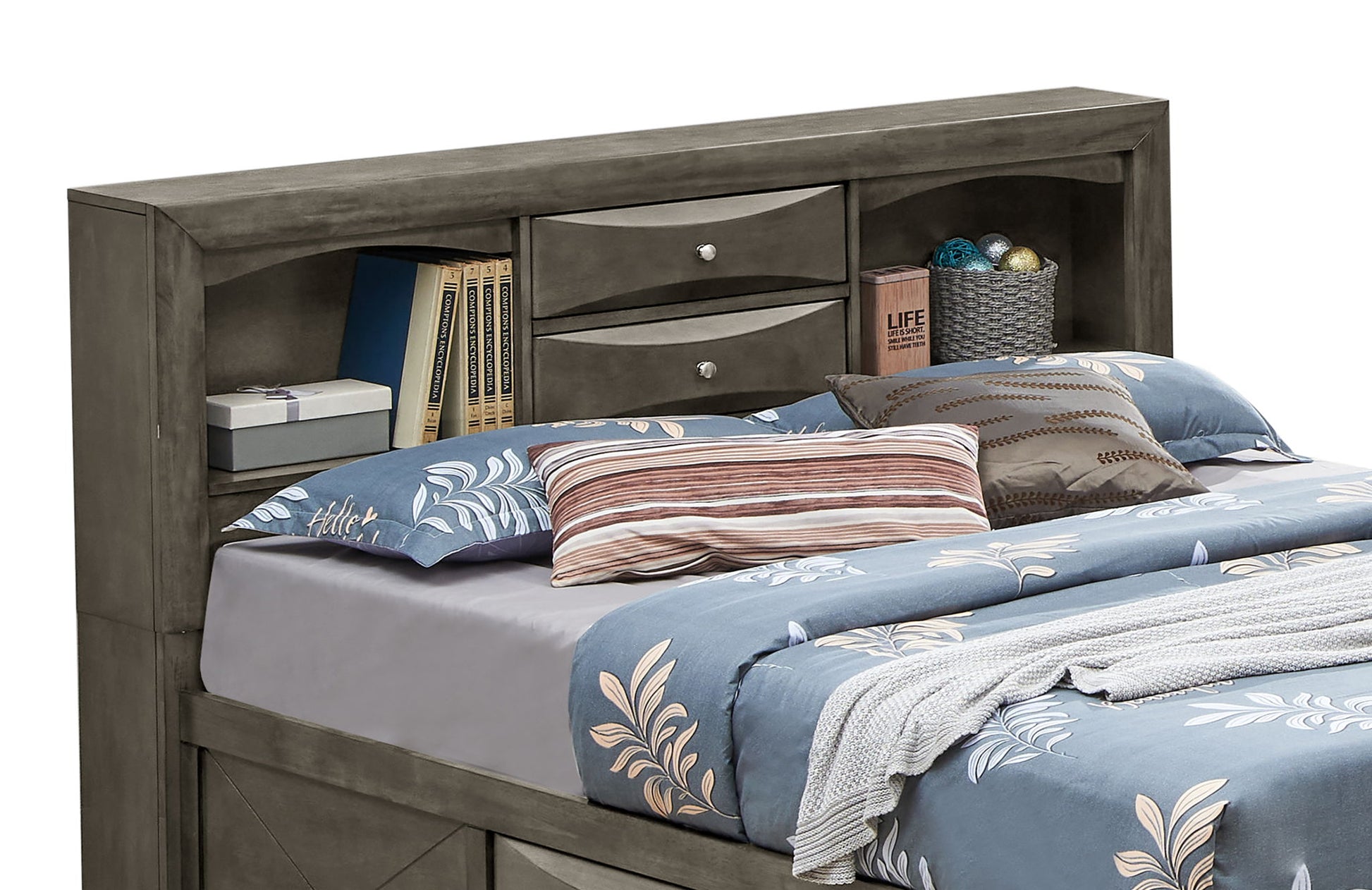 The Marilla Storage Bed With Bookcase Headboard in gray features shelves, drawers, a box, and a basket. It’s styled with blue and gray bedding, patterned pillows, and a folded blanket at the foot.