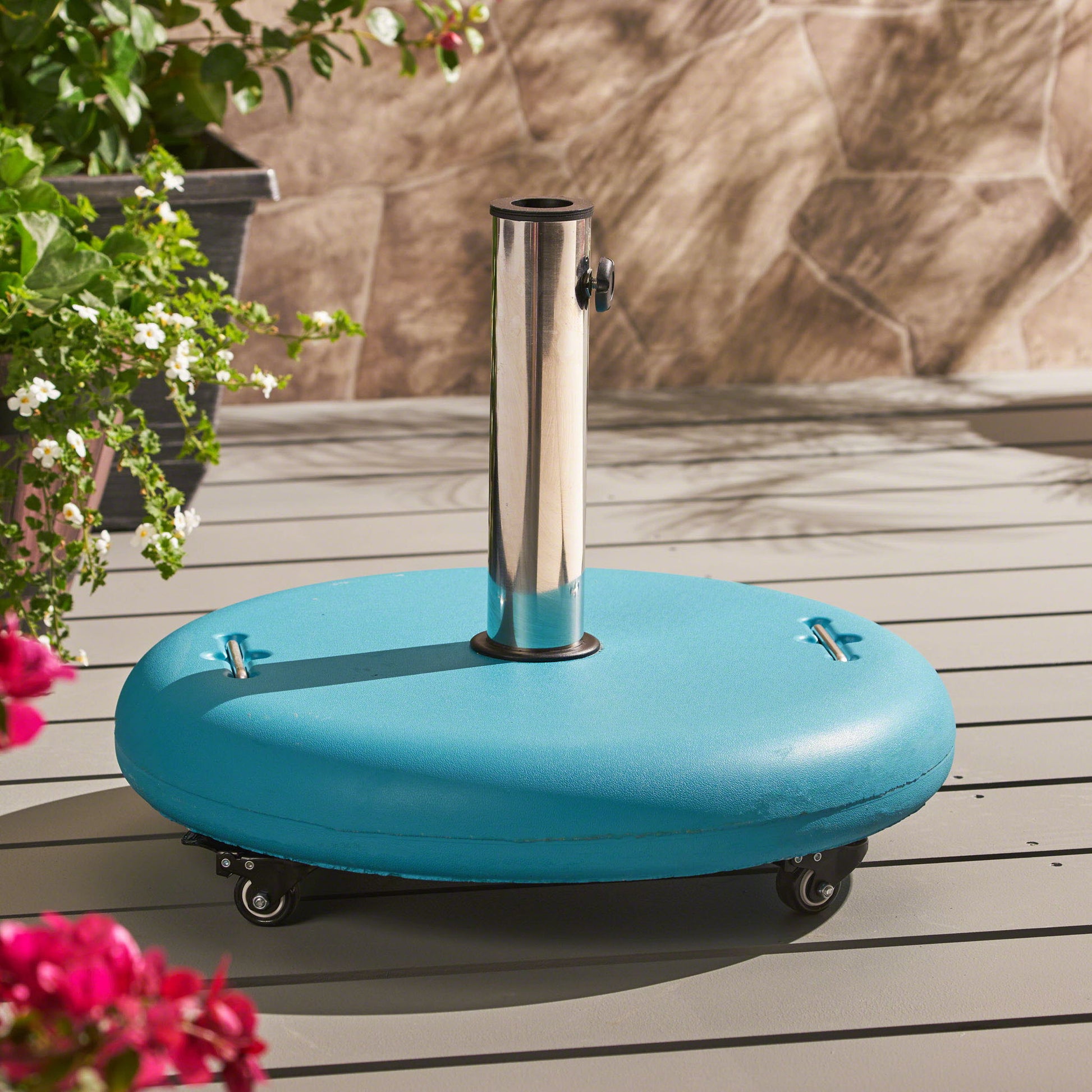 The Wheelie Round Umbrella Base with Stainless Steel Holder stands on a gray deck. Its blue, round design with wheels is complemented by nearby green plants and flowers, as sunlight creates soft shadows on the ground.