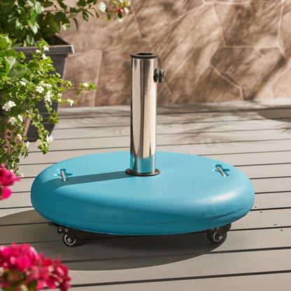 The Wheelie Round Umbrella Base with Stainless Steel Holder stands on a gray deck. Its blue, round design with wheels is complemented by nearby green plants and flowers, as sunlight creates soft shadows on the ground.