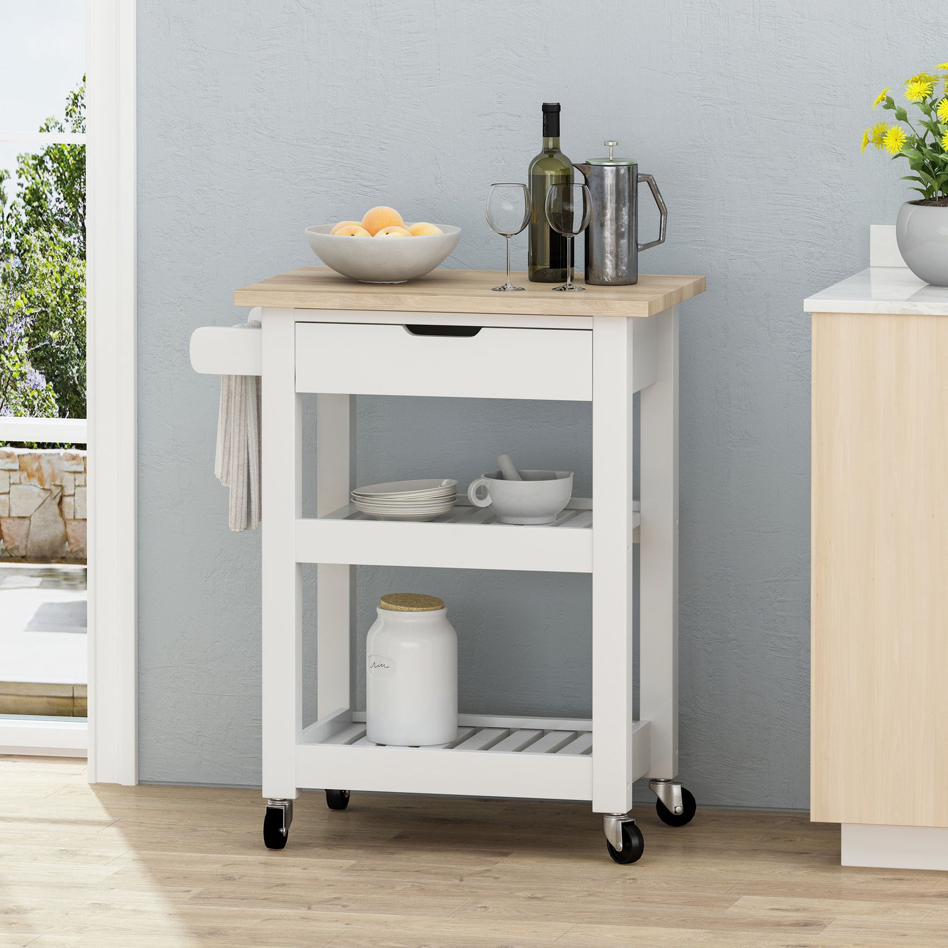 The Tattnall Kitchen Cart With Wheels features a wooden top, white body, drawer, two shelves, and black wheels. It holds a fruit bowl, two wine glasses, coffee pot, and jar. A towel hangs on the side; yellow flowers sit nearby on the counter.