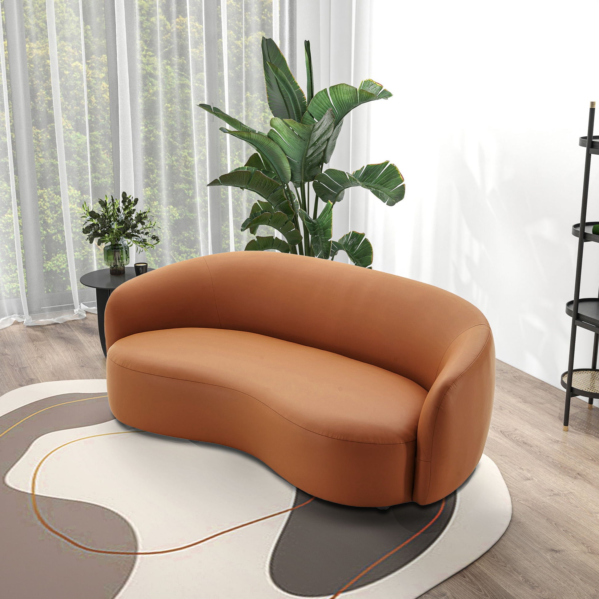 The Porter - Comfortable Sofa, with its modern curved brown leather design, sits on a patterned rug in a bright room with wood floors, beside a large green plant and a side table with flowers by sheer-curtained windows.